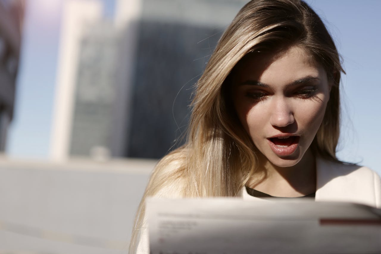 A young woman with blonde hair expresses surprise while reading a newspaper outdoors in daylight.